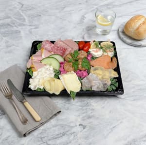 En välfylld tallrik i en svart plastbricka, upplagd på en bädd av grönkål.  Vänster sida: Skivad skinka, salami, leverpastej, krämig potatissallad och gurkskivor.  Mitten: Fyra köttbullar på rosa rödbetssallad, samt en bit brieost och en rullad skiva hårdost.  Höger sida: Gravad lax, två sorters sill (löksill och senapssill) samt ägghalvor med räkor, citron och dill.  Toppen: Två små körsbärstomater som garnering.  Kort sagt: En klassisk kall tallrik med alla de vanligaste favoriterna.