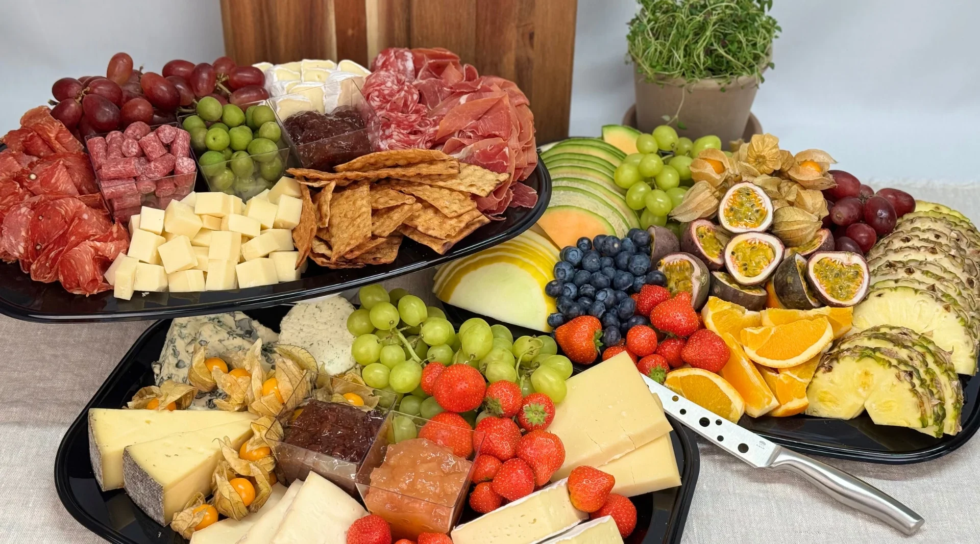 Ett bord är dukat med två stora fat fyllda med olika ostar, charkuterier, kex och färsk frukt. På det övre fatet finns bland annat druvor, oliver, salami, brieost, lufttorkad skinka, kex och osttärningar. På det undre fatet syns ett urval av hårdostar, mögelost, färska jordgubbar, blåbär, gröna druvor, physalis och två sorters marmelad. Brevid ligger färska melonskivor, apelsinklyftor, passionsfrukt och ananasskivor. I bakgrunden stå en kruka med färska örter. En ostkniv ligger på ena fatet.