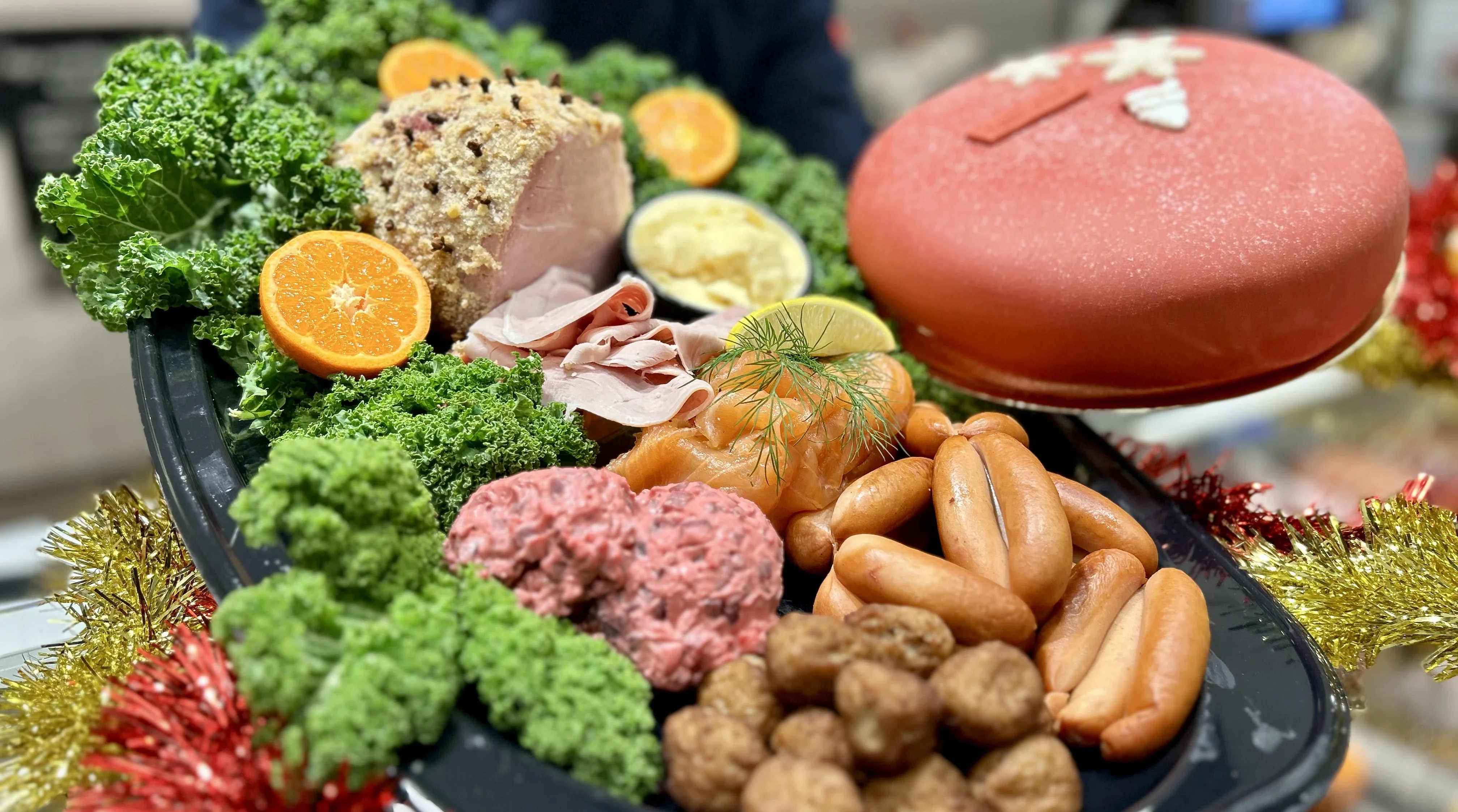 Ett stort fat med prinskorv. julskinka, köttbullar och en röd prisesstårta med julmotiv dekorerat med grönkål och apelsin