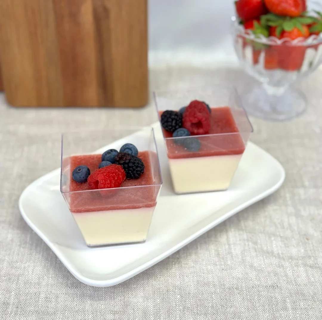 Två glas med pannacotta toppad med bärsås, hallon, blåbär och björnbär, serverade på ett vitt fat.