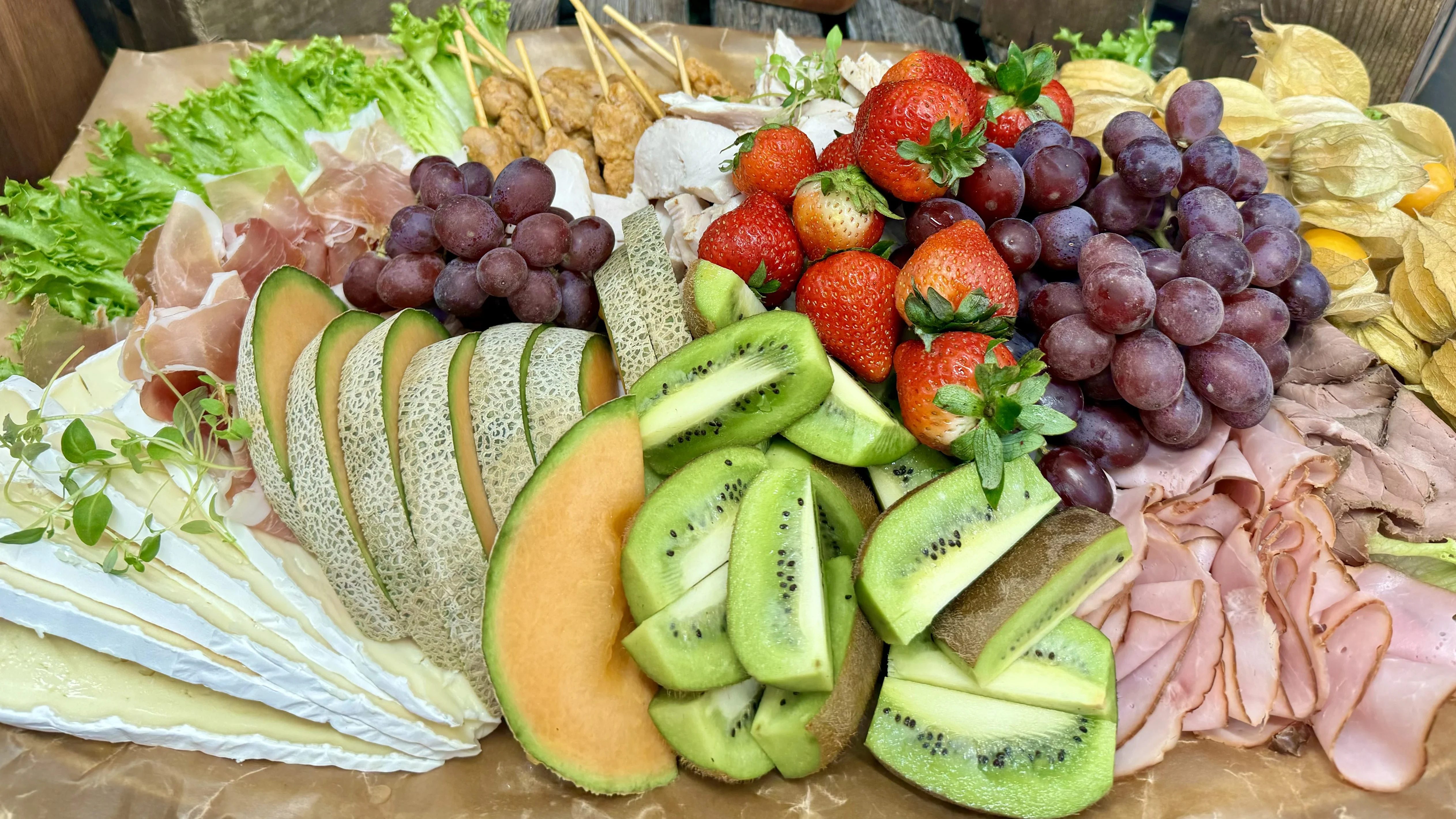 En solplanka fylld med olika charkprodukter, kycklingspett, frukt och grönsaker