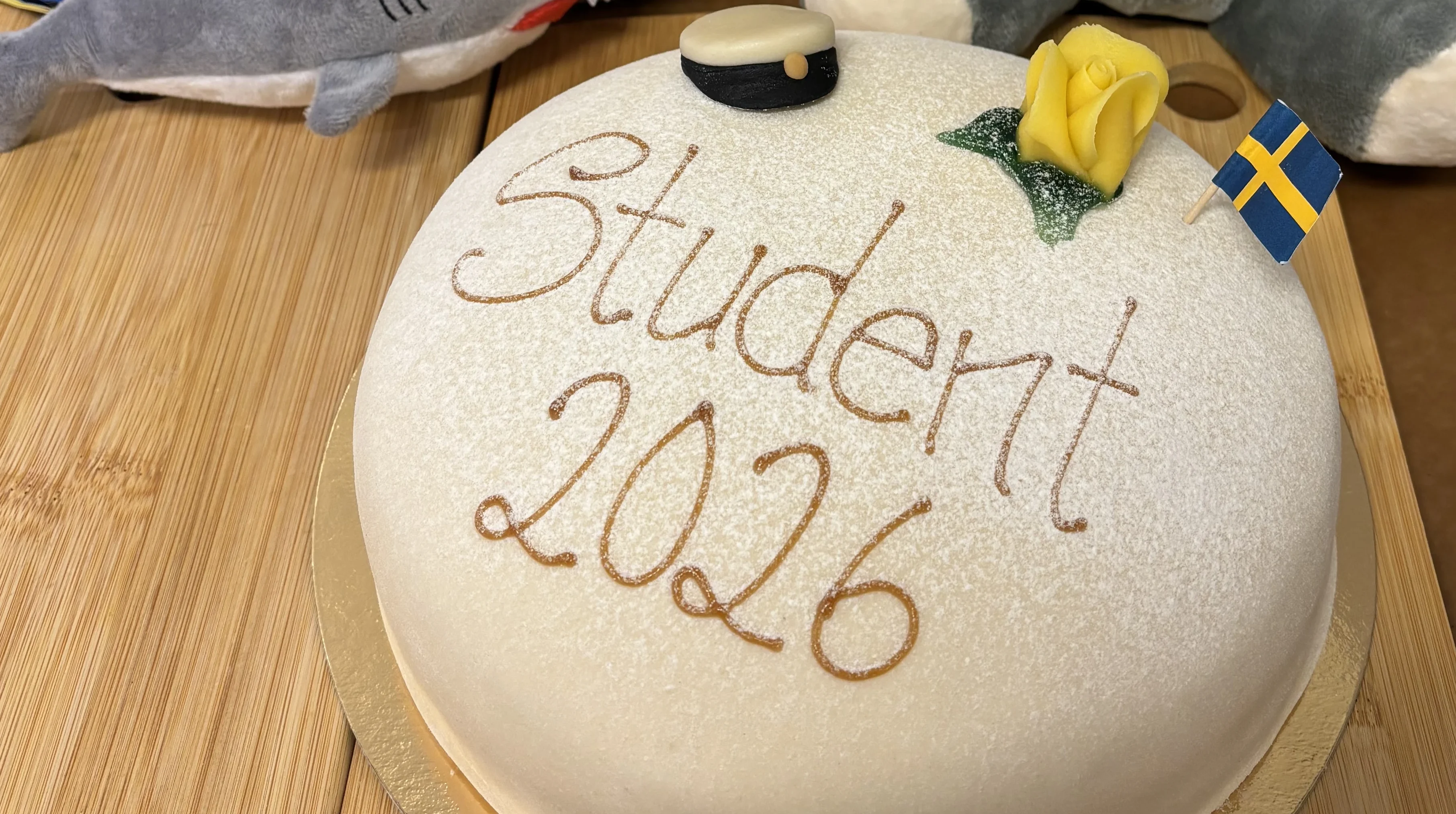 en rund vit prinsesstårta med texten studenten 2026. gul marsipanros och en liten ätbar studentmössa