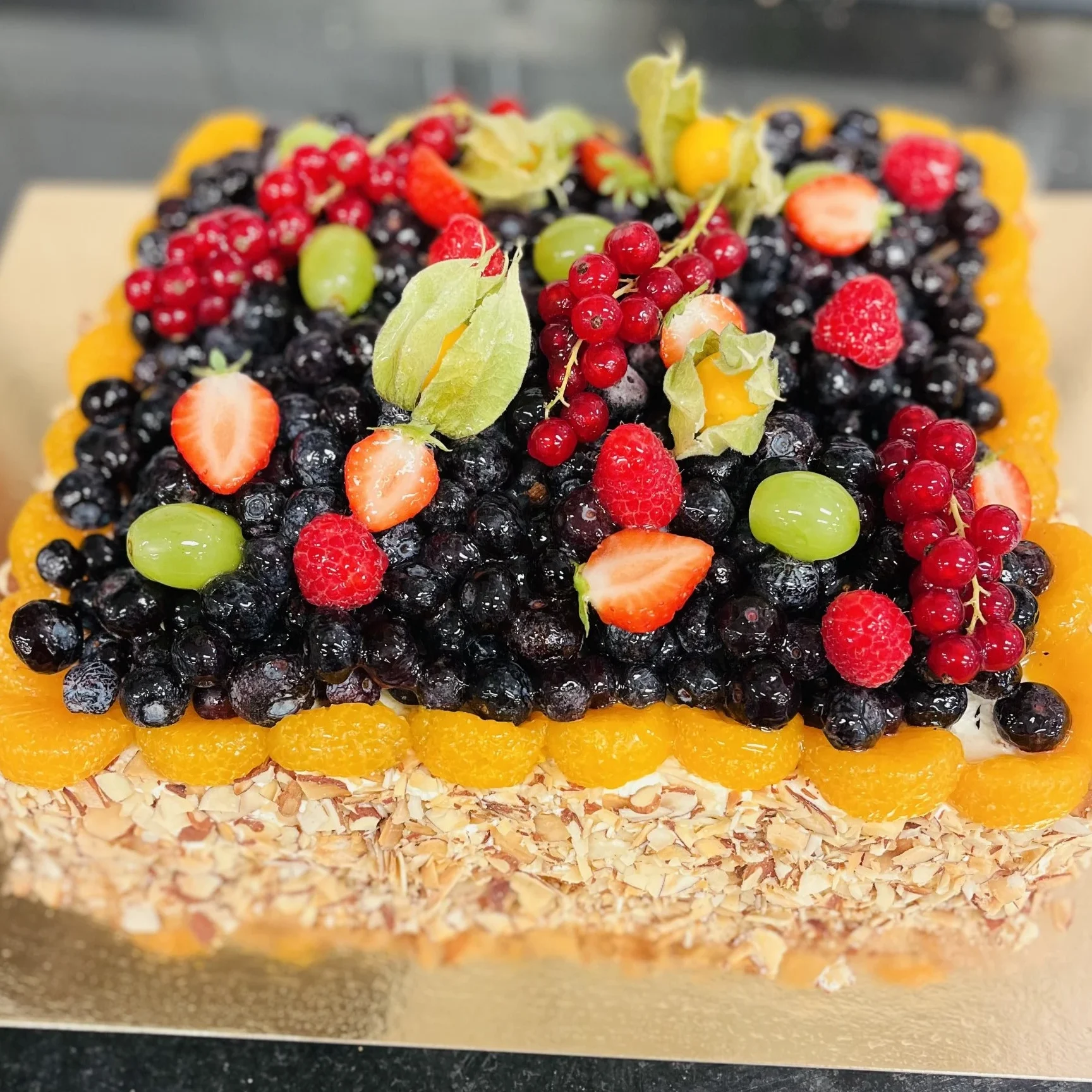 Fyrkantig frukttårta med grädde, täckt med mandelspån på sidorna och toppad med blåbär, hallon, jordgubbar, röda vinbär, physalis, vindruvor och mandarinklyftor.