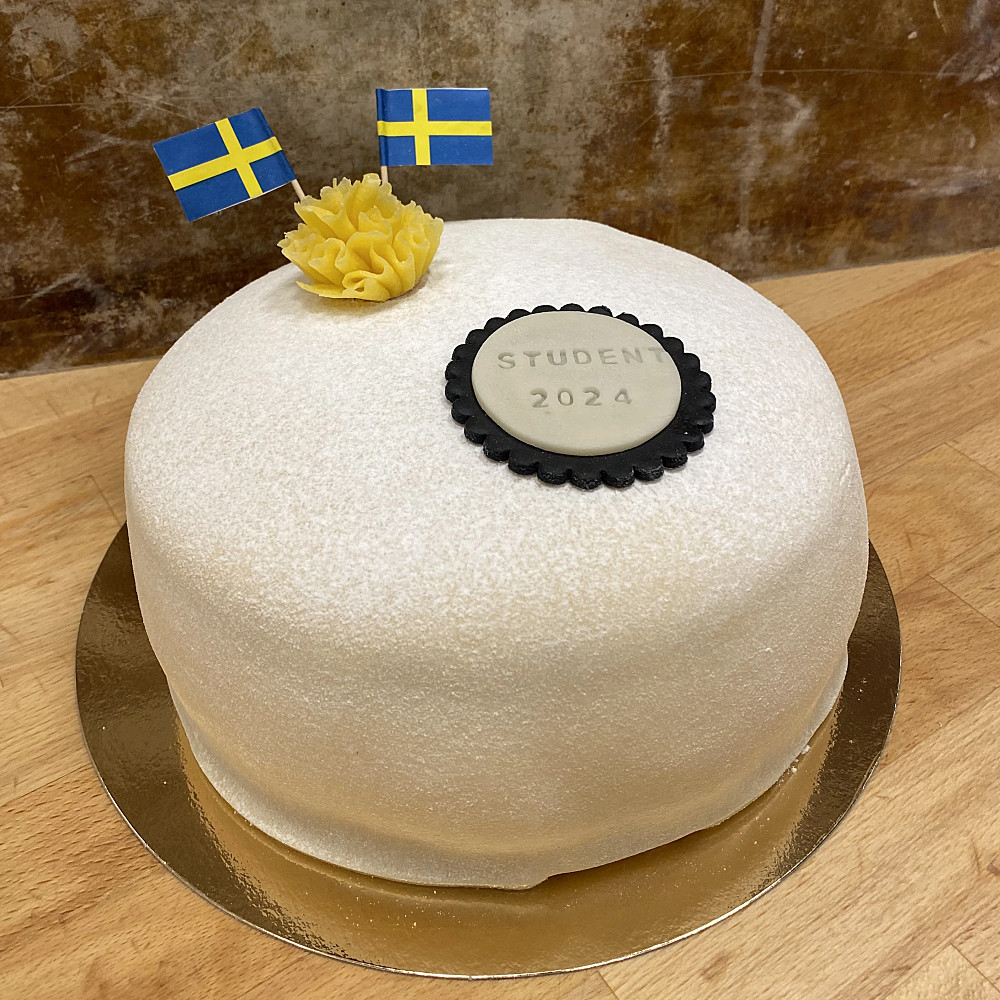 Vit prinsesstårta dekorerad med svenska flaggor, gul blomma och text - student.