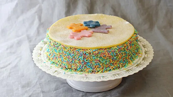 En lekfull tårta på sockerkaksbotten med fyllning av hallonmousse och vaniljkräm. Dekorerad med massor av blandat strössel på kanterna. Ovansida av marsipan med dekorpusselbitar.