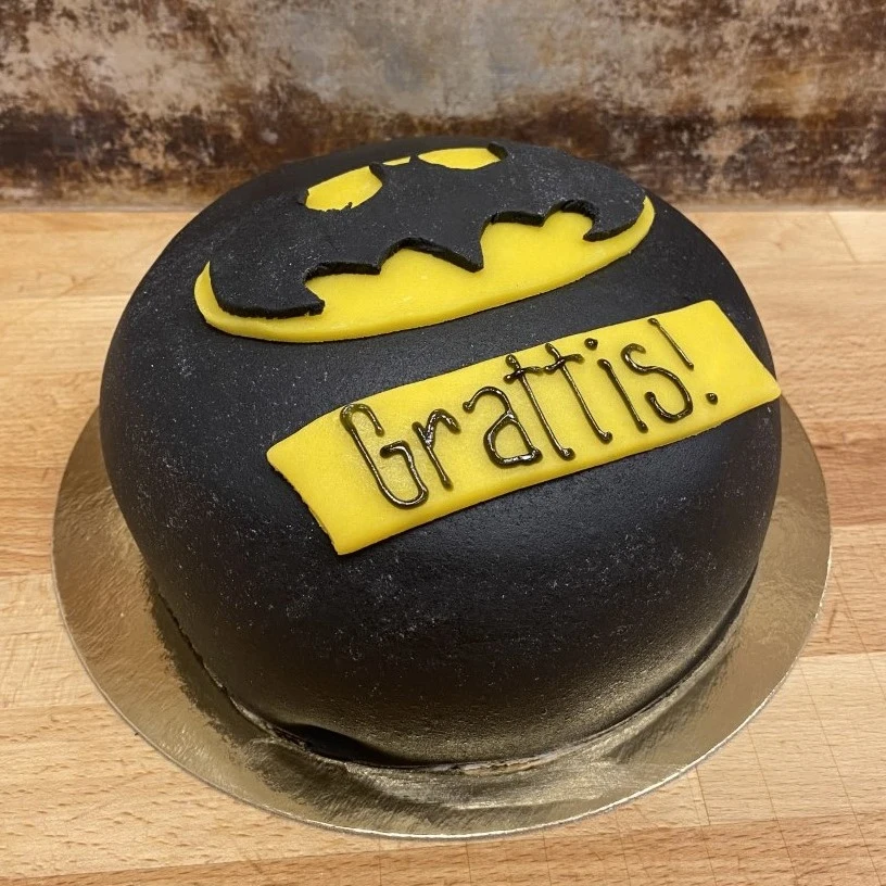 En tårta med svart marsipan, batman-märke i gult och svart och en gul marsipanbit med text/namn.