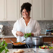 Vardagskocken står i ett kök och håller i en kvist basilika bredvid en stekpanna med mat.