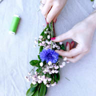 En person arrangerar blommor och gröna blad på ett tyg bredvid en trådrulle.