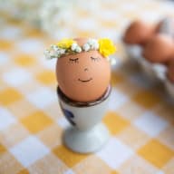Ett ägg med tecknat ansikte och blomsterkrans sitter i en äggkopp på en rutig duk.