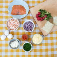 En bild visar olika ingredienser utlagda på en rutig duk, inklusive lax, räkor, rödlök, ägg, bröd och grönsaker.