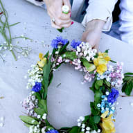 En person håller en blomsterkrans gjord av olika blommor och gröna blad på en betongyta.