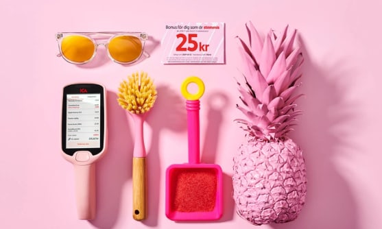 En bild på ett självscanningshandtag, en bonuscheck, solglasögon, diskborste, ananas i rosa på rosa bakgrund.