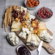 Festligt ostfat med blandade ostar, frukt, brödpinnar samt marmelad och tomater vid sidan.