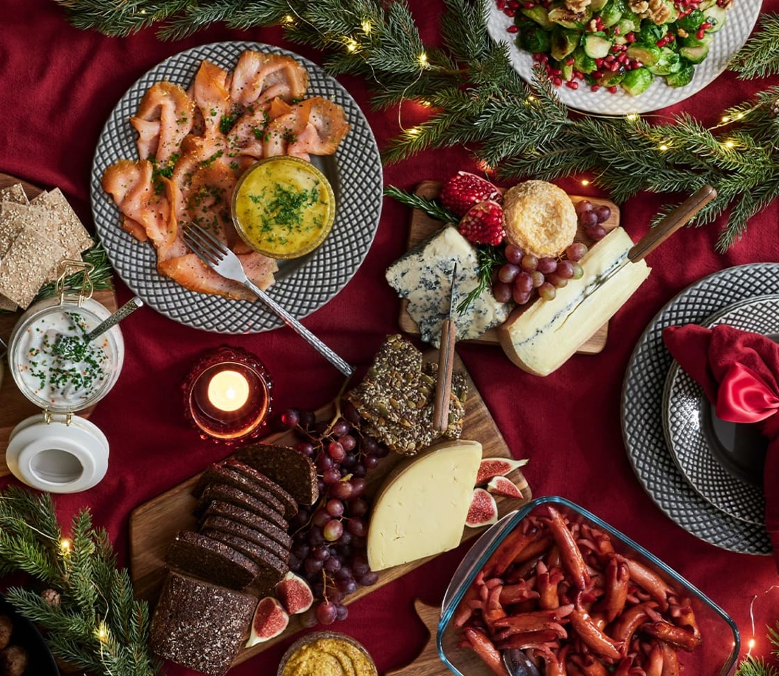Uppdukat julbord med ostbrickor, bröd, kallrökt lax, småkorvar och tillbehör runt ett tänt ljus.