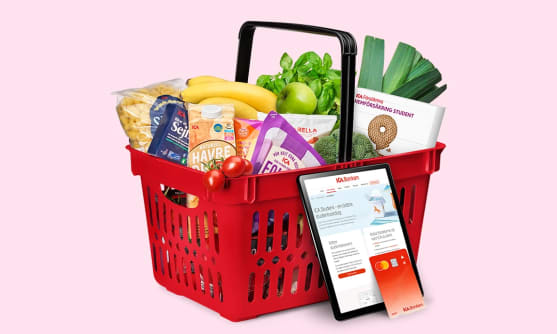 En röd korg fylld med ICA-varor mot en rosa bakgrund Men en surfplatta och ett ICA-bankkort framför.