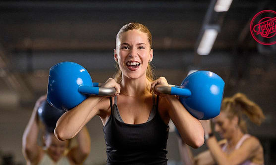 En glad tjej tränar med två blå kettle bells på en Friskis & Svettis-anläggning.