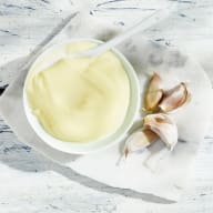 Skål med krämig sås (aioli) och en sked bredvid några vitlöksklyftor på en marmorskiva.