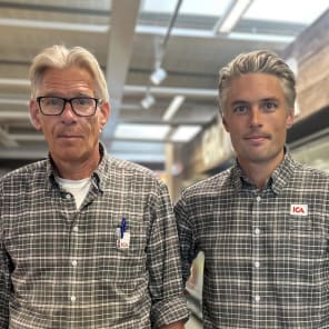 Två ICA‑anställda i matchande rutiga skjortor står bredvid frukt- och grönsaksdisken i en mataffär.