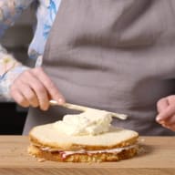 En person breder smör på en brödskiva på en skärbräda, med en skål och ett saltkar bredvid på en köksbänk.
