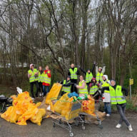 En grupp personer i reflexvästar står vid en hög med fyllda soppåsar och kundvagnar i en skogsomgivning.