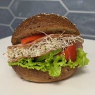 En rund smörgås fylld med salladsblad, skinka, skivad tomat, gurka och groddar.