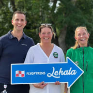 En man och två kvinnor står på rad och tittar leeende in i kameran. Kvinnan i mitten håller en blå pil där det står "Flygfyrens lokala".