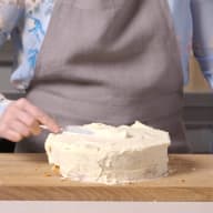 En person sprider frosting på en tårta på en skärbräda i ett kök.