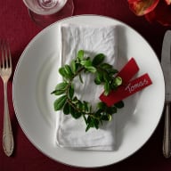 Ett dukat bord med en ett glas, en tallrik med en vit tygservett med en minikrans av lingonris och en röd papperslapp med namn på som placeringskort