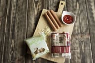 En träskärbräda med korvar, ketchup, en påse potatischips och en förpackning med köttprodukter på ett träbord.