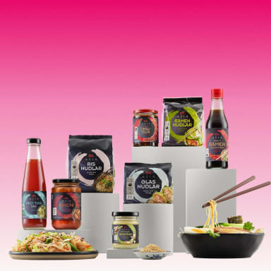 En samling matprodukter står på vita podier bredvid en skål med nudlar och en tallrik med mat.