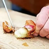 En person skalar en vitlöksklyfta med en kniv på en träskärbräda.