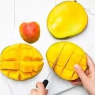 En person skär en mango med en kniv bredvid en hel mango och ett äpple på en vit yta.