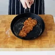 En person lägger ett stycke panerat kött i en stekpanna på en skärbräda.
