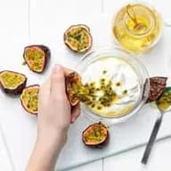 En person håller en halv passionsfrukt över en skål med yoghurt, omgiven av fler passionsfrukter och en flaska honung.
