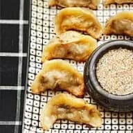 Fyra dumplings ligger på en rutig yta bredvid en skål med sesamfrön.