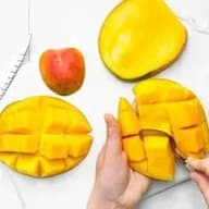 En person skär upp en mango bredvid ett helt äpple på ett bord.