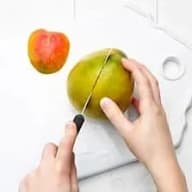En person skär en frukt med en kniv på en skärbräda medan en annan frukt ligger bredvid.