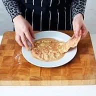 En person viker en tunn pannkaka på en vit tallrik som står på en träskärbräda.