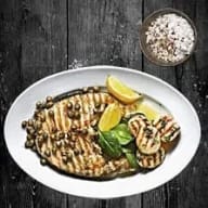 En tallrik med grillad fisk och citronskivor står på ett träbord bredvid en skål med kryddor.