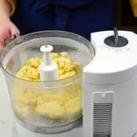 En person använder en matberedare med gula ingredienser i behållaren bredvid en vit apparat på en yta.