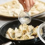 En person häller vätska från ett litet glas över dumplings i en skål, med en tallrik synlig i bakgrunden.