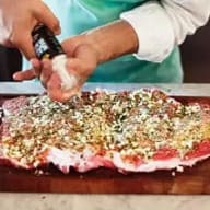 En person kryddar ett stort köttstycke på en skärbräda med en kryddflaska.
