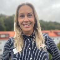 Person i randig skjorta med namnbricka står på en balkong med bostadshus och träd i bakgrunden.