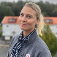 En person i ICA-jacka står på en balkong med bostadshus och träd i bakgrunden.