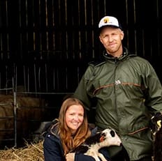 En person sitter på en stol och håller en hund medan en annan person står bredvid i ett stall med får och halm på golvet.