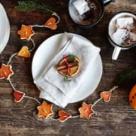 En tallrik med en tygservett. På servetten ligger en torkad apelsinskiva med en liten granris-bukett och kanelstång fäst mitt på apelsinskivan med ett rött band. Runt tallriken ligger en girlang avtorkat apelsinskal.