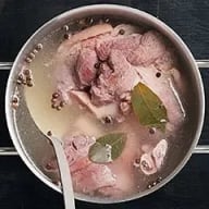 En skål med köttsoppa innehållande köttstycken, lagerblad och små runda kryddor i klar buljong.