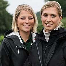Två personer i mörka jackor håller en trälåda fylld med jordgubbar framför en odling.