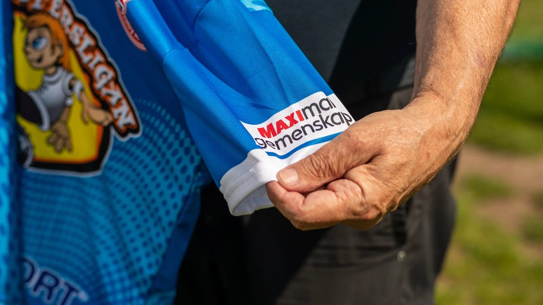 På bilden syns en tröjan från Kvartersligen och på ärmen finns loggan Maximal Gemenskap, en äldre person håller i tröjan och sträcker ut ärmen så att loggan syns tydligt.