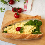 Baguette fylld med äggsallad och bladgrönt, toppad med en körsbärstomat.